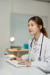 Young asian female doctor wearing a lab coat and stethoscope is typing on a computer keyboard in her brightly lit office, focused on her work