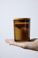 Person holding a candle in hand, white background with brown candle in glass jar with wooden lid