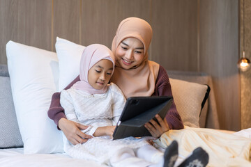 Muslim mother and daughter wearing hijabs using tablet together in bedroom bonding and enjoying quality time, showing technology usage for learning, education and entertainment, modern muslim family