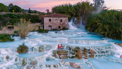 a diverse couple of man and woman enjoy the hot spring thermal waters at Saturnia in Tuscany, Italy, as the sunrise casting a warm glow over the stunning landscape and cascading pools. 