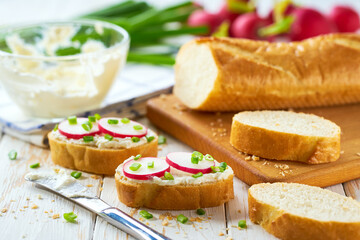 Appetizing crunchy baguette slices with cream cheese and radish slices and freshly chopped chives on a white wooden table.