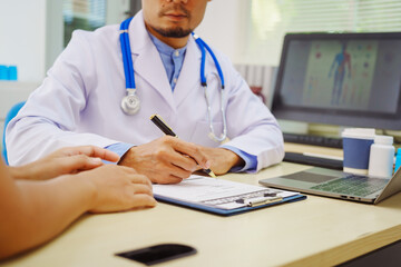 A male doctor sits at a desk in a hospital,reviewing a female patient’s medical history.He...