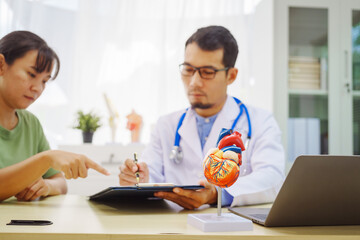 Fototapeta premium A doctor man sits at a desk in a hospital, explaining heart disease symptoms to a female patient. They discuss chest pain, palpitations, fatigue, dizziness, and the risks of myocardial ischemia