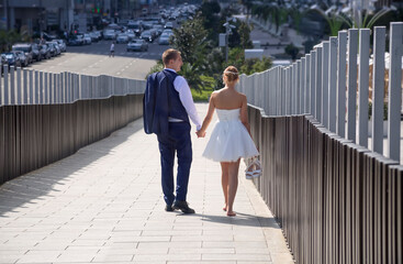 The newlyweds, tired and happy, walk down the bridge in a modern city on a hot summer day, leaving the camera