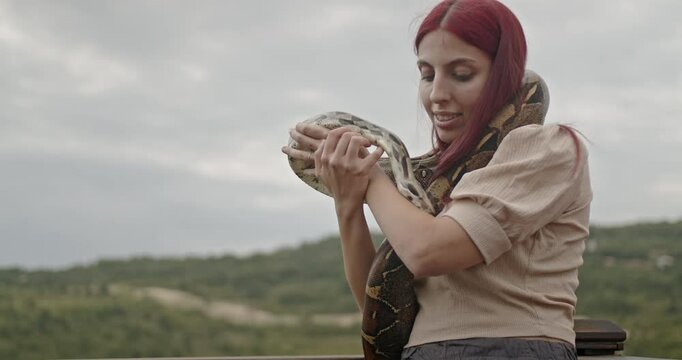 A happy young woman with red hair plays with her pet snake outdoors on a cloudy day.