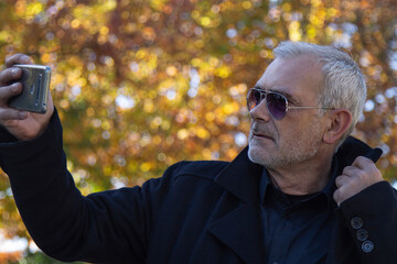 Mature man using mobile phone in the park in autumn for taking a selfie or live video