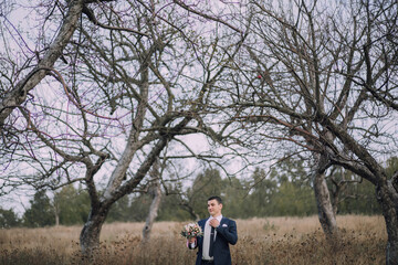 A man in a suit stands in a field of trees, holding a bouquet of flowers. The scene is serene and peaceful, with the man looking out into the distance