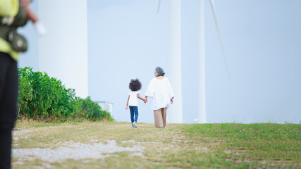 An Asian grandmother walks hand in hand with her grandchild near a wind turbine, reflecting...