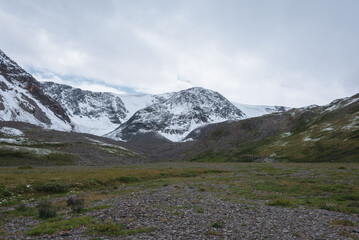 Dramatic cold landscape with green valley against snow covered large mountain range under rainy cloudy gray sky. Gloomy rainy weather in high mountains. Alpine view to big snowy mount in rain time.