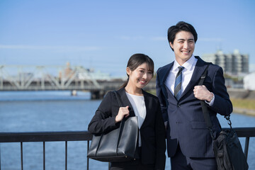 Two freshmen, a man and a woman, who look like young new salesmen standing by the river in suits.