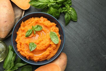 Tasty mashed sweet potato with basil in bowl and fresh vegetables on dark gray textured table, flat lay. Space for text