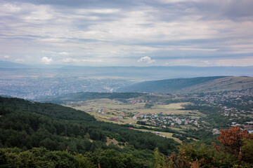 Summer mountain view of Tbilisi, Georgia. Soft clouds cast gentle light over the city and its green landscape, with lush trees in the foreground, creating a calm, scenic atmosphere