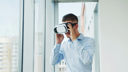 Man Using Virtual Reality Headset in Modern Office. A young man in a light blue shirt explores virtual reality using a VR headset in a bright, modern office with large windows.