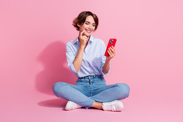 Full size portrait of lovely cheerful girl sit floor finger touch chin use smart phone isolated on...
