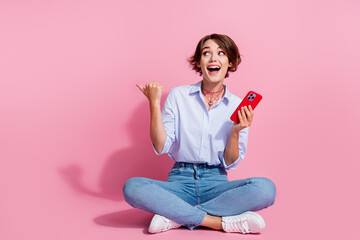 Full body portrait of charming impressed lady sit floor hold smart phone look indicate finger empty space isolated on pink color background