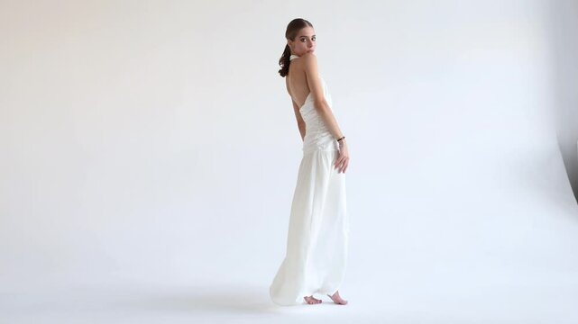 Young fashion model posing in long stylish Flowing white dress against isolated white background in studio. Woman beauty and grace