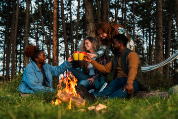 Friends making a sunset toast at forest campsite. Natural moment of joy as diverse group raises...