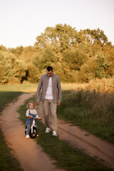 Obraz premium Father teaches a young child to ride a balance bike in the park in the summer. The concept of fatherhood and family weekend outings.