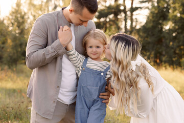 Fototapeta premium Happy Caucasian family of three, man, woman and child happy in the field in summer. Casual clothes. Mom blonde with long hair in a white dress.