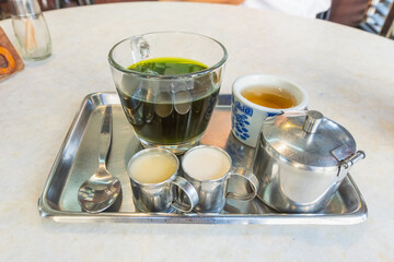 A refreshing green beverage served in a glass, accompanied by two small cups and a milk container on a metal tray, perfect for tea time.
