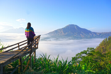Fototapeta premium Woman standing alone from behind on a wooden bridge, looking at the view of clear blue sky over mountains with fog in the morning. Inspirational motivational background. Mount Batur, Kintamani, Bali
