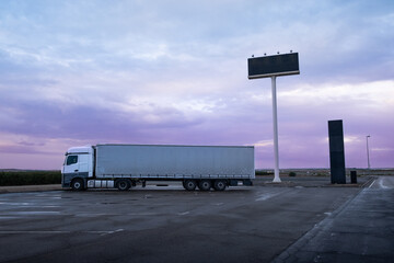 A big white truck parked at a service station at dusk to spend the night. The asphalt ground is wet from the rain. Concept of rest and safety in the transportation of goods by road © LaMorenita