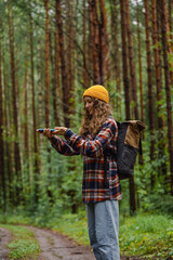 Young female hiker with backpack and yellow beanie using smartphone for navigation while exploring lush green forest trail, enjoying peaceful nature and outdoor adventure