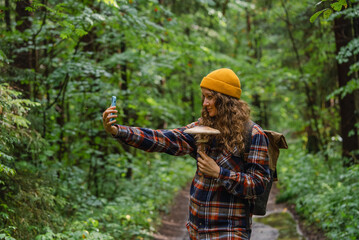Fototapeta premium Young biologist or hiker woman wearing yellow beanie and plaid shirt taking a selfie with a mushroom she found in a green forest during a summer hike