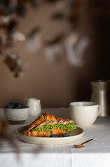 Croissant sandwich with ham, cheese and lettuce served on a white plate. Breakfast with croissant, cup of coffee and blueberries. Vertical orientation.