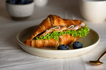Croissant sandwich with ham, cheese and lettuce, with cup of coffee and blueberries on a table.