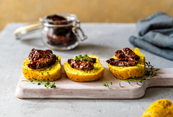 Slices of turmeric bread topped with sun-dried tomatoes and garnished with fresh thyme on a light wooden cutting board, with a jar of sun-dried tomatoes in the background