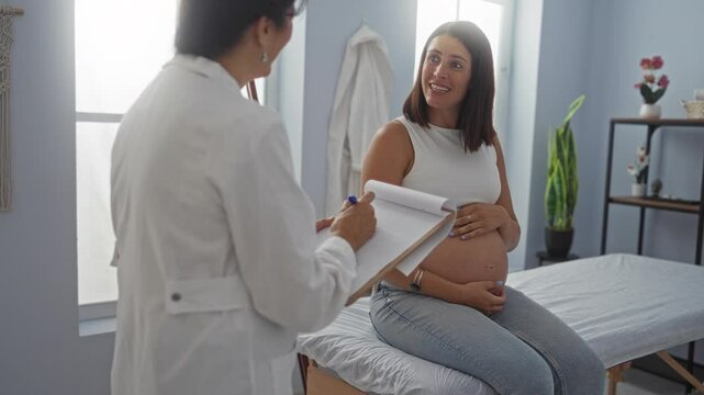 Pregnant woman and female doctor in a clinic discussing medical notes during a prenatal checkup with bright indoor setting.