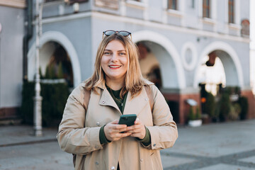 Happy cheerful young woman with backpack walking on city street checks her smartphone. Portrait of...