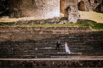 Bride and groom hold hands on ancient stone steps against a backdrop of historic ruins, creating a romantic and timeless scene.