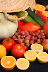 Round straw bag and various seasonal fruits and vegetables on dark background. Summer and fall produce. Selective focus.