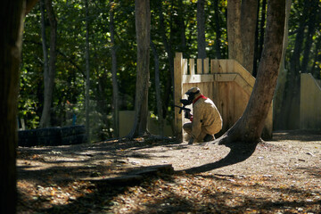 Paintball player in protective mask aims gun, takes cover under fire, paint splashes. Paintball player hide behind wooden barricade, shooting paintballs. Paintball players.
