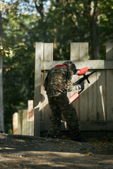 Fototapeta premium Paintball player in protective mask aims gun, takes cover under fire, paint splashes. Paintball player hide behind wooden barricade, shooting paintballs. Paintball players.