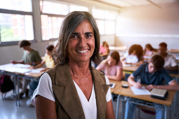 Portrait of happy mature Caucasian female teacher at high school posing in class with confidence. Middle-aged tutor woman looking smiling at camera, classroom with unfocused teenage students at desk