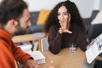 A woman in a casual outfit gestures as she explains her ideas to a man sitting across from her. The two are engaged in a collaborative session, surrounded by books and work materials on a wooden desk