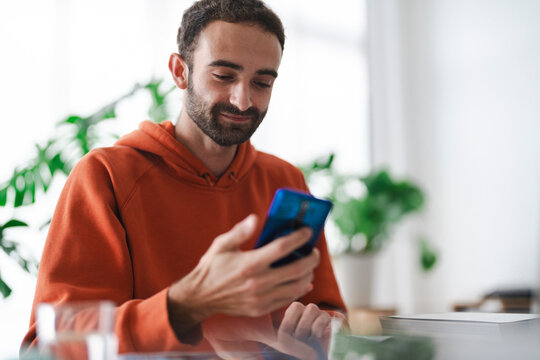 man in an orange hoodie looks at his smartphone while sitting at a desk. He appears focused, with books and a glass in the foreground, creating a cozy and productive atmosphere with natural lighting
