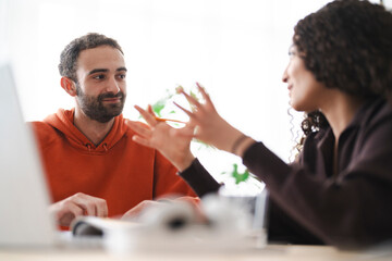 man and a woman engage in an intense discussion, possibly brainstorming or problem-solving. They sit across from each other with a laptop and books, indicating a collaborative or creative environment.