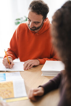 A man in an orange hoodie writes in a notebook at a table, surrounded by books and papers, fully engaged in a focused learning environment.