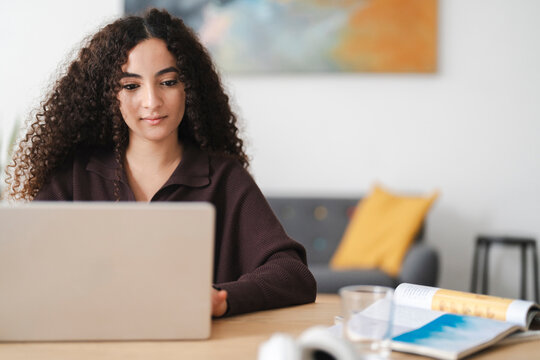 A young Arab woman with curly hair is deeply focused as she types on her laptop, creating a quiet and studious atmosphere at the table.