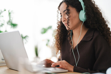 A young woman with curly hair wearing teal headphones intently works on her laptop, surrounded by plants, conveying a productive and modern remote work environment.
