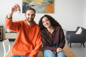 A cheerful young couple sitting together at a table, with the man in an orange hoodie holding up a set of keys, symbolizing a fresh beginning or new home in a brightly lit, modern room.