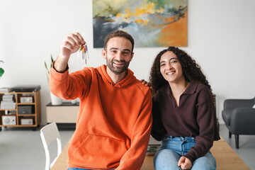 A cheerful young couple sitting together at a table, with the man in an orange hoodie holding up a set of keys, symbolizing a fresh beginning or new home in a brightly lit, modern room.