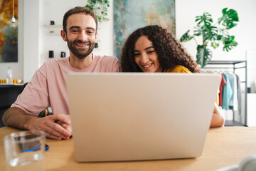 A happy couple seated together, looking at a laptop screen in a warm, cozy room decorated with plants, enjoying shared moments and comfortable companionship.