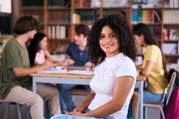 Portrait happy young Latin female student sitting in library with classmates unfocused in background. Group friends meeting together in study room. High school girl poses looking smiling at camera