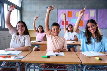Portrait of happy group diverse elementary school students sitting in classroom with hands raised....