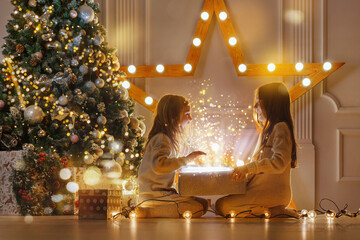 Happy sibling girls opening christmas presents. Children with gift boxes and christmas star. Decorated living room with traditional fire place.